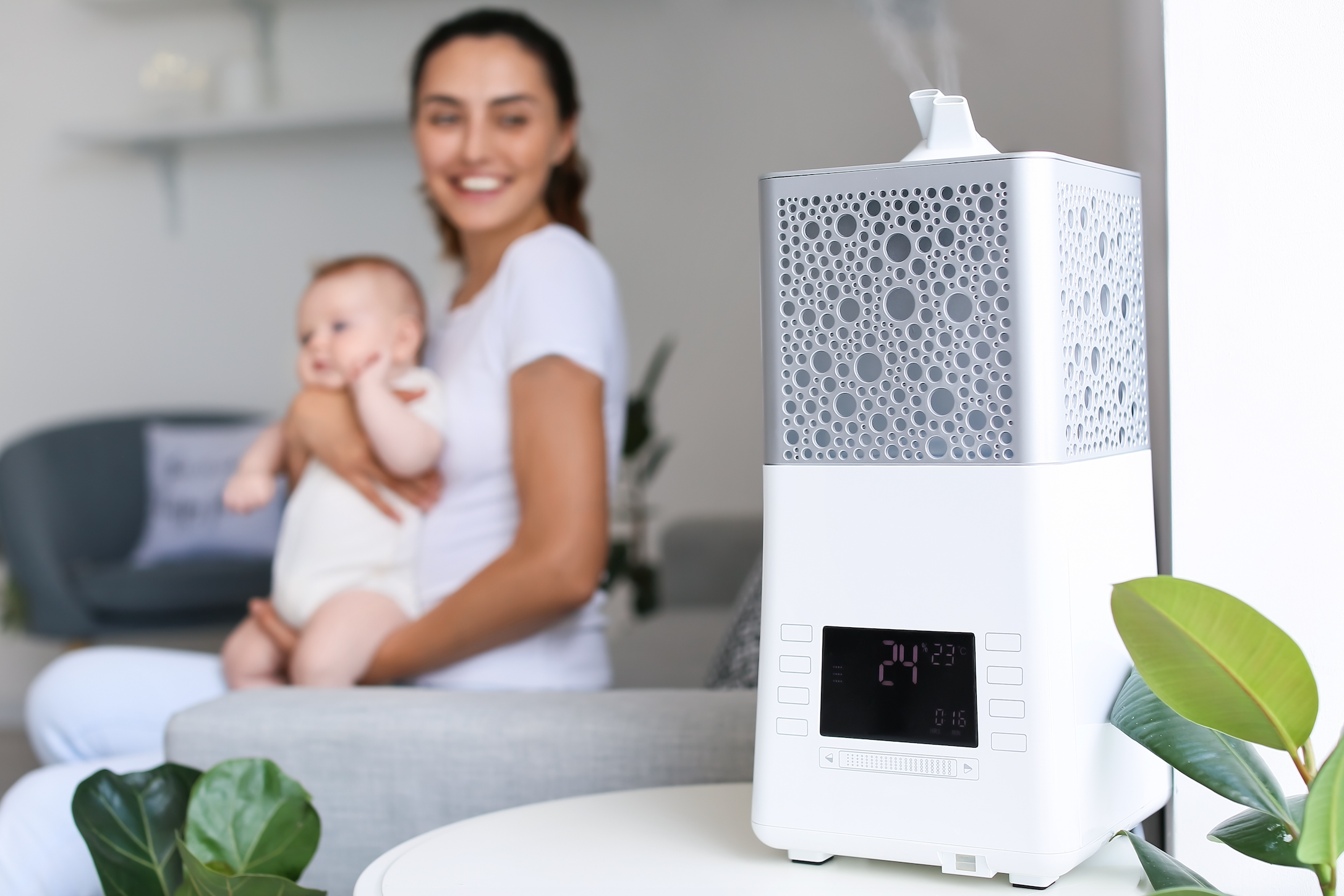 Smiling mother holding toddler on couch, looking over at humidifier 