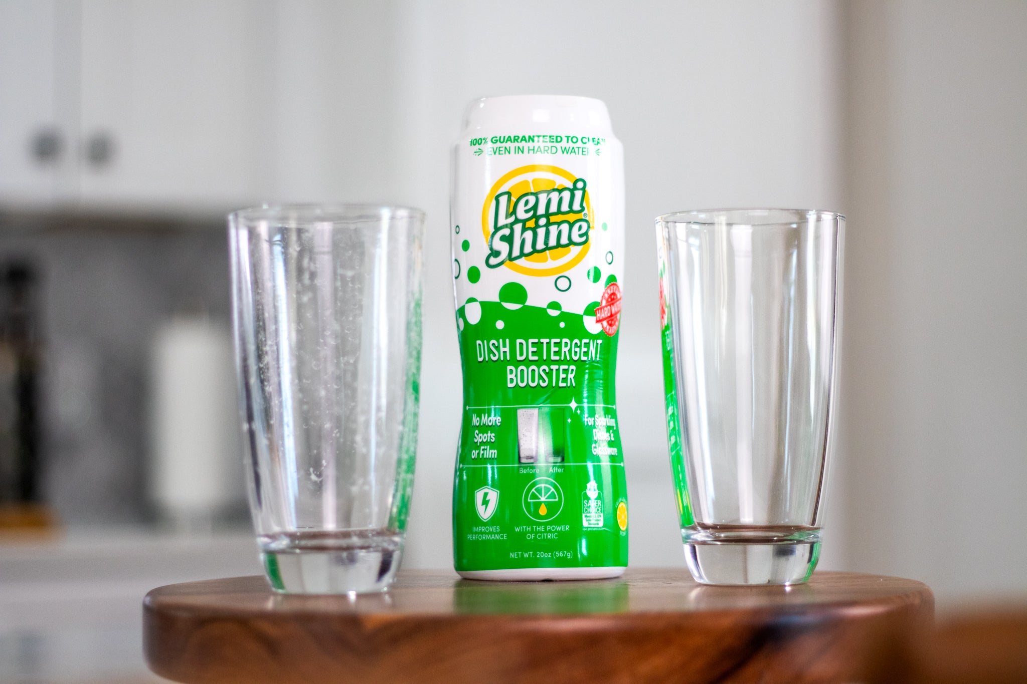 Lemi Shine dish detergent booster bottle between two glasses on a wooden surface.